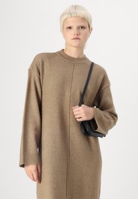 Robe pull marron tricotée avec des manches longues, un col côtelé et des coutures latérales ; le modèle tient un petit sac noir. Texture lisse, coupe décontractée.