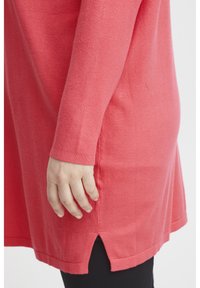 Tunic a maglia corallo con maniche lunghe e spacchi laterali all'orlo. Texture morbida, vestibilità comoda e scollatura rotonda. Leggings neri indossati sotto.