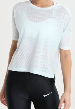 Frau trägt ein weißes, perforiertes Kurzarmtop und schwarze Nike-Leggings mit weißem Swoosh-Logo, steht mit entspannten Armen.