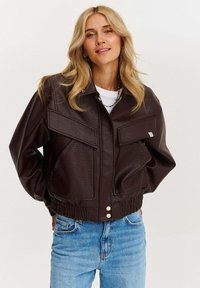 Jachetă bomber din piele ecologică maro, cu două buzunare pe piept, talie elastică și închidere cu fermoar. Finisaj texturat și siluetă lejeră.