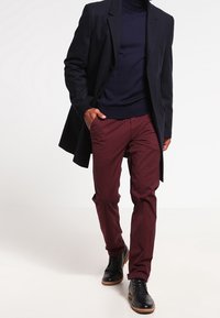 Marineblauwe jas over een donkere coltrui, gecombineerd met kastanjebruine broek en zwarte schoenen met beige zolen. Gladde stof en een getailleerde pasvorm.