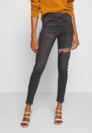 Jean skinny noir avec un détail déchiré au genou, fabriqué en denim extensible. Associé avec des sandales à talons striées blanches et un pull moutarde.
