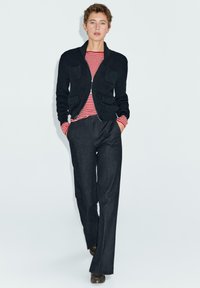 Black ribbed knit cardigan with two front pockets, over a red and white striped long-sleeve shirt and dark tailored trousers.