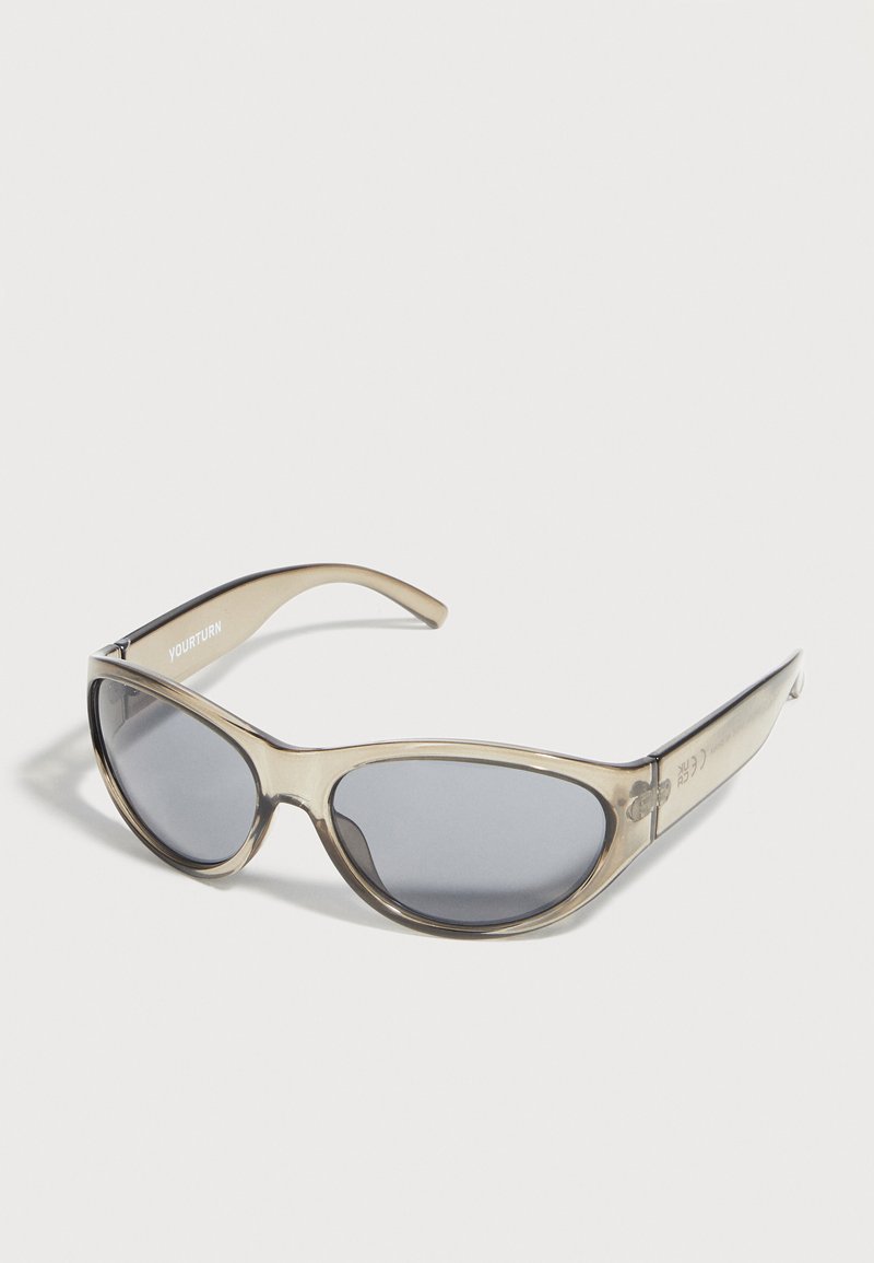 Lunettes de soleil beige transparentes à forme œil de chat avec des verres teintés foncés posées sur une surface blanche.