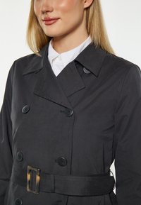 Czarny, podwójnie zapinany płaszcz trench z paskiem w talii, dużymi guzikami i kołnierzem. Posiada gładką, matową teksturę oraz dopasowany krój.