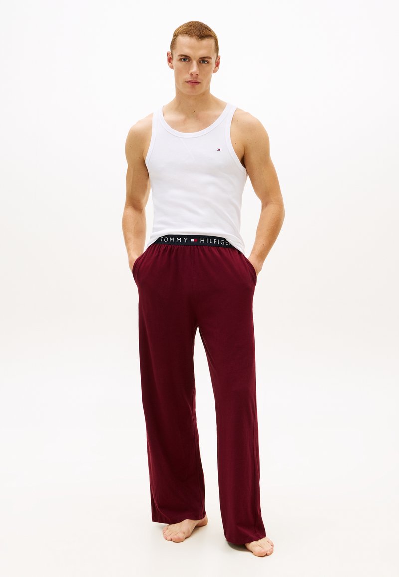 Jeune homme aux cheveux courts portant un débardeur blanc et un pantalon rouge ample, les mains dans les poches, debout pieds nus sur un fond uni.