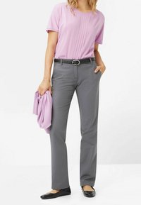 Top corto a maniche corte di un leggero viola con pieghe verticali, abbinato a pantaloni grigi e una cintura nera. La modella tiene un maglione leggermente viola. Ballerine nere.