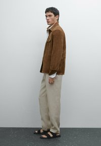 Brun mockajacka med två bröstfickor, beige vida byxor och svarta platta sandaler. Tyget verkar mjukt med en matt finish.