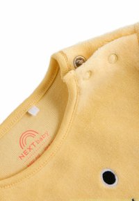 Gelbes Fleece-Babybekleidungsstück mit Rundhalsausschnitt, ausgestattet mit Druckknöpfen und einem schwarzen gestickten Detail. Weicher Stoff, mit Etikett NEXTBaby.