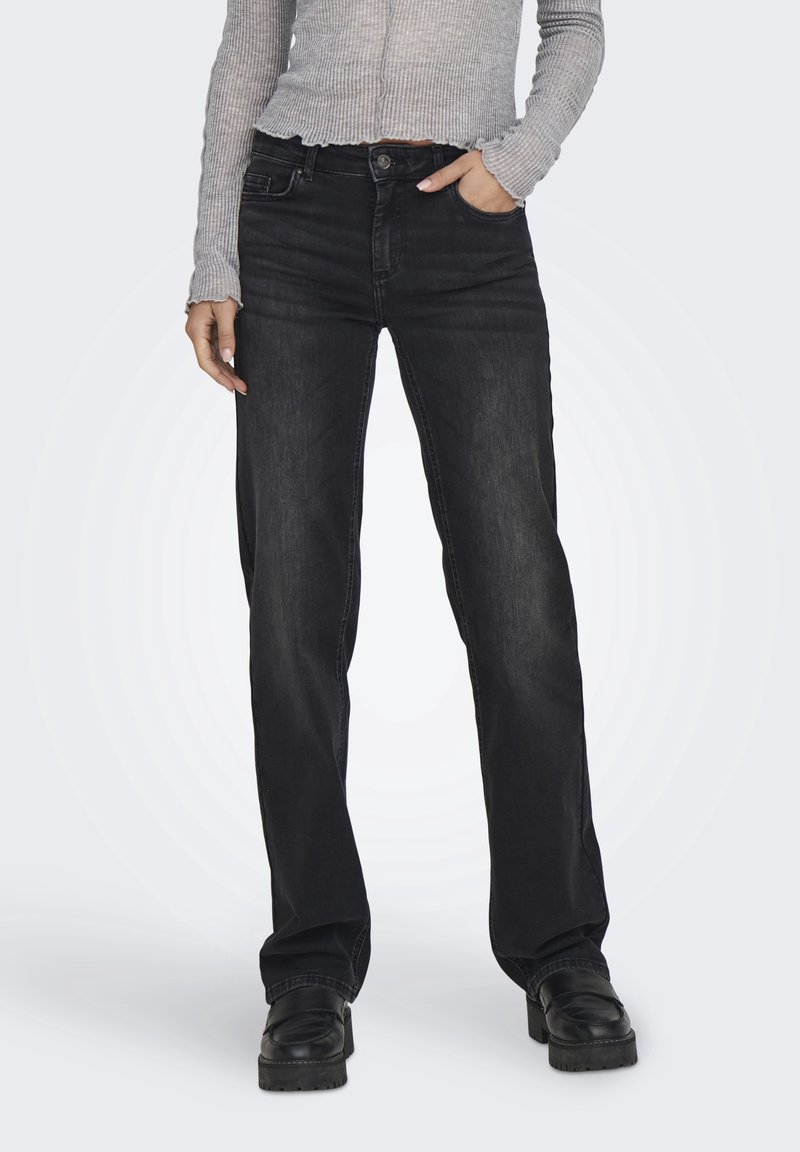 Femme portant un jean noir taille haute droit, un haut gris côtelé à manches longues et des chaussures plateformes noires, main dans la poche, debout.
