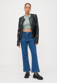 Νεαρή γυναίκα με σκουλαρίκια κρίκους, φορώντας μια μαύρη δερμάτινη ζακέτα, μίντ πράσινο crop top, μπλε καμπάνα τζιν και μαύρες λόφες.