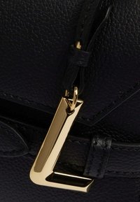 Sac en cuir texturé noir avec un charm en métal doré en forme de "L" bien en vue et des finitions assorties, présentant un design épuré et minimaliste.