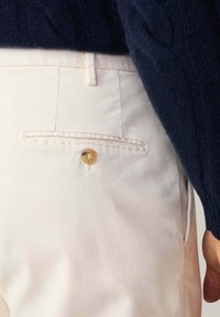 Lichtgekleurde broek met een achterzak, voorzien van een ronde gouden knop. De stof lijkt glad en is gedeeltelijk bedekt door een donkerblauwe trui.