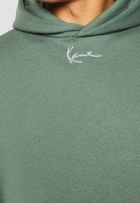 Gros plan sur un sweat à capuche vert avec une petite signature blanche brodée près du col et une partie du cou visible au-dessus du vêtement.