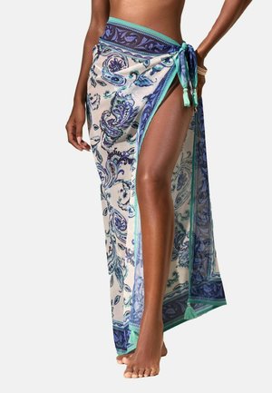 Frau trägt einen transparenten, blau-türkis gemusterten Sarong, der an der Hüfte gebunden ist und ein Bein mit nackten Füßen vor einem schlichten Hintergrund zeigt.