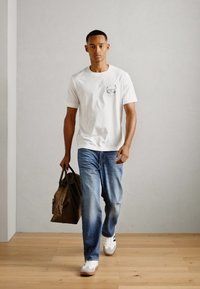 Tricou alb cu mânecă scurtă și buzunar, blugi albaștri și adidași albi. Poartă o geantă de umăr din material maro. Podea din lemn, fundal deschis.