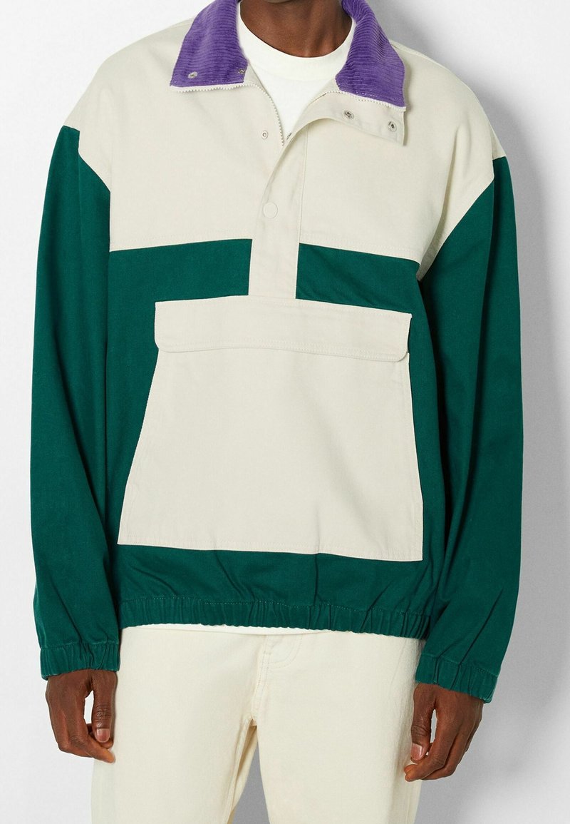 Homme portant une veste-pull vert et beige avec une grande poche avant et un col en velours côtelé violet, associée à un pantalon beige.