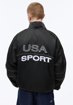 Person mit kurzen Haaren, die eine schwarze Jacke mit der Rückseitenstickerei "USA SPORT" trägt, kombiniert mit blauen Jeans, die wegschaut.