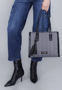 Borsa a spalla a spina di pesce nera e grigia con dettagli in pelle, manici superiori e un dettaglio con nappina. Abbinata a pantaloni in denim e stivali neri a punta.