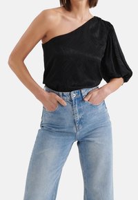 Schwarzes einseitiges Oberteil aus strukturiertem Stoff mit Puffärmel-Detail, kombiniert mit hellblauen, hoch taillierten Schlagjeans.