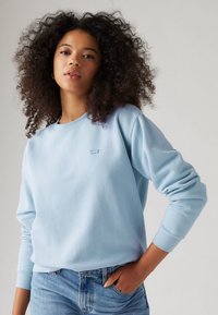 Sweat-shirt bleu clair en polaire douce, avec un col rond et des poignets côtelés. Petit logo brodé sur la poitrine, associé à un jean en denim.
