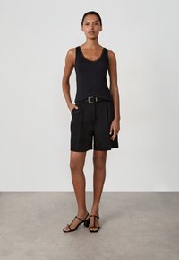 Top nero senza maniche con scollatura a zig-zag, abbinato a pantaloni corti neri su misura con cintura. Il modello indossa sandali neri con tacco.
