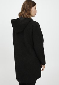 Fekete kapucnis kabát texturált anyagból, amely bolyhos megjelenésű. Lazán illeszkedik és a comb közepéig ér.