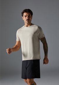 T-shirt de sport beige clair avec des manches courtes et des accents texturés, porté avec un short noir ; un modèle masculin dans une pose dynamique.