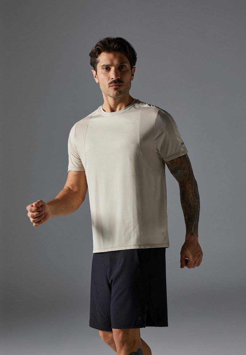 T-shirt de sport beige clair avec des manches courtes et des accents texturés, porté avec un short noir ; un modèle masculin dans une pose dynamique.