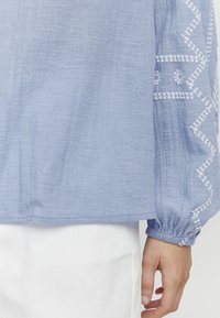 Vaaleansininen, teksturoitu pitkähiha-paita, jossa valkoiset kirjailut hihojen varrella ja rypytetty hihansuu ranteessa.