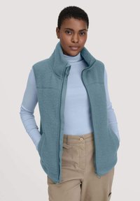 Hellblaues Fleece-Weste mit hohem Kragen, Frontreißverschluss und zwei Seiten Taschen. Getragen über einem hellblauen Langarmshirt und beigen Hosen.