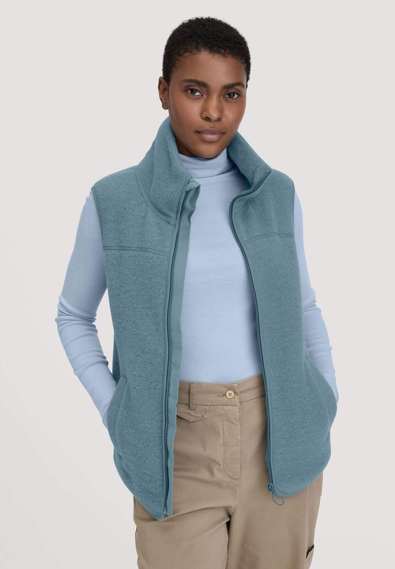 Hellblaues Fleece-Weste mit hohem Kragen, Frontreißverschluss und zwei Seiten Taschen. Getragen über einem hellblauen Langarmshirt und beigen Hosen.