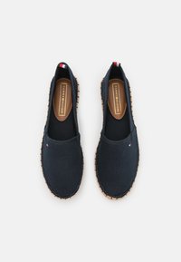 Baskets slip-on en toile marine avec une semelle en jute tressé, présentant un petit accent rouge, blanc et bleu au niveau du talon et une semelle intérieure marron.