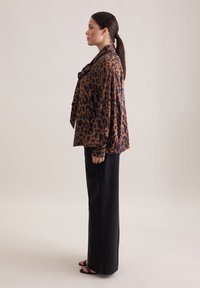Blusa in raso con stampa leopardata in marrone e nero, con una vestibilità ampia, dettaglio con fiocco sul collo e maniche lunghe e fluide, abbinata a jeans neri.