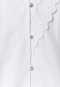 Blusa bianca con colletto in pizzo ondulato, cinque bottoni lucidi sul davanti e tessuto di cotone liscio con una finitura croccante.
