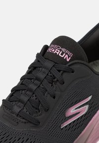 Black athletic shoe with mesh upper, pink and silver accents, cushioned sole, and eyelet lacing system. Features Skechers branding on tongue.