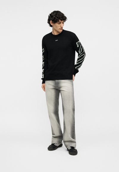 Νεαρός άνδρας με σγουρά μαλλιά, ντυμένος με μαύρο πουλόβερ με λευκά σχέδια στα μανίκια, ξεθωριασμένο γκρι τζιν και μαύρα αθλητικά παπούτσια, στέκεται μπροστά σε λευκό φόντο.