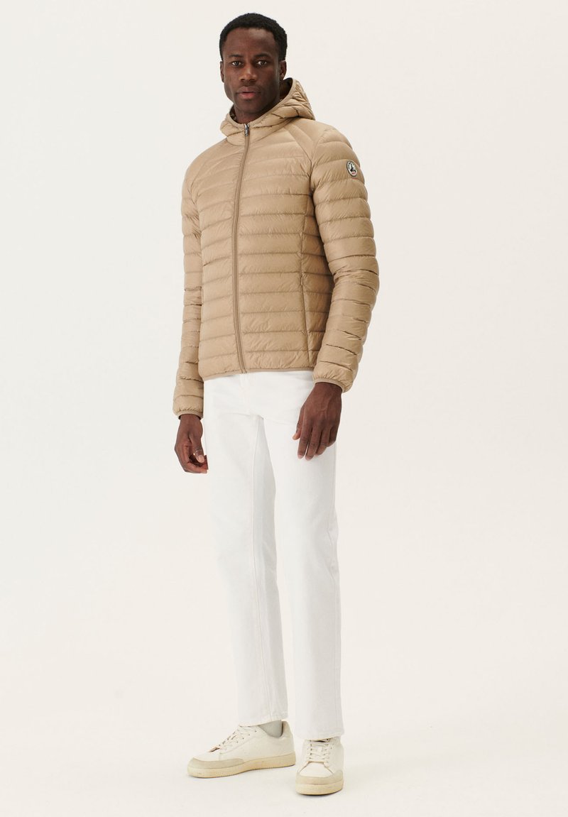 Beige pufferjacka med huva, quiltad yta, frontdragkedja och logodetalj. Matchad med vita byxor och beige sneakers.