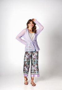 Jasnofioletowy sweter z dzianiny z wiązaniem w talii. Spodnie w tropikalny wzór z liści w kolorze turkusowym, brązowym i fioletowym. Sandały na paski w kolorze brązowym.