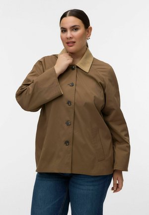 Kobieta o ciemnych włosach nosi brązową kurtkę na guziki z kołnierzem ze sztruksu i niebieskie dżinsy, pozuje z ręką przy kołnierzu na białym tle.