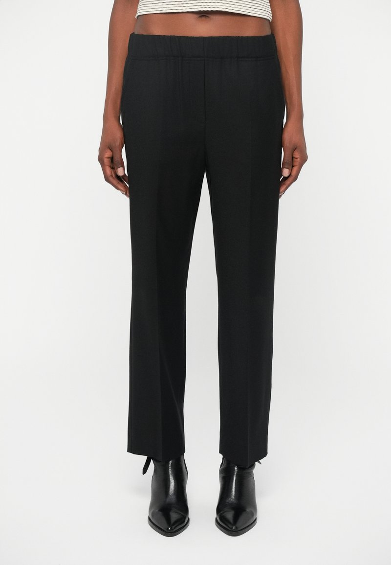 Calças largas pretas, com uma cintura elástica, textura suave e um corte ajustado. Usadas com botas de salto alto pretas até o tornozelo.