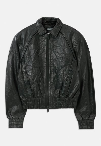 Must selledega kortsus must nahktagi, millel on ees lukk, elastsed varrukad ja alläär, ning külgmised kaldu taskud, valge tausta peal.