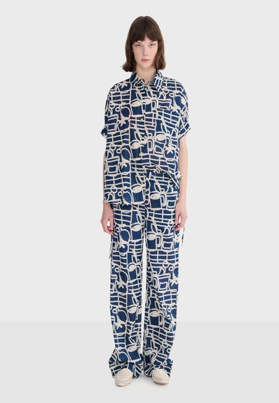Femme portant une chemise et un pantalon assortis amples avec un motif géométrique et de fruits bleu marine et blanc, chaussée de chaussures blanches tissées, debout devant un fond uni.