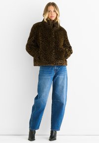 Giacca in pelliccia ecologica con stampa leopardata marrone, con colletto alto e chiusura frontale a bottoni, abbinata a jeans azzurri a gamba larga e stivaletti neri.