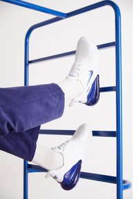 Vitaid sportskor med en texturerad ovandel och en klarblå häl. Bärs med vita strumpor och marinblå byxor, framför en blå metallram.