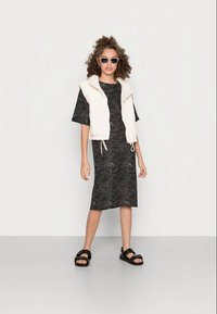 Colete acolchoado branco sobre um vestido preto com padrão, de mangas curtas. Sandálias pretas e óculos de sol completam o visual. Texturas suaves visíveis.