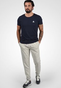 Dunkelblaue Baumwoll-T-Shirt mit einem kleinen weißen Dreieckslogo, kombiniert mit hellgrauen Jogginghosen und dunklen Sneakers mit weißen Schnürsenkeln.