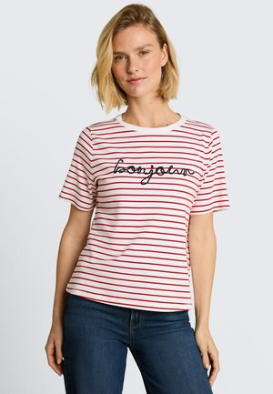 Femme aux cheveux blonds courts porte un t-shirt blanc à rayures horizontales rouges avec le texte "bonjour", associé à un jean bleu foncé.