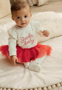 Hvid skjorte med flæser, røde prikker og teksten "Santa Baby". Rød tylskørt med glimmerdetaljer. Sidder på en blød overflade.