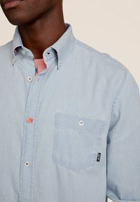 Lichtblauwe button-up shirt van denimstof, met een borstzak, contrasterende knoopkleuren en een patroon op de binnenrand van de kraag.